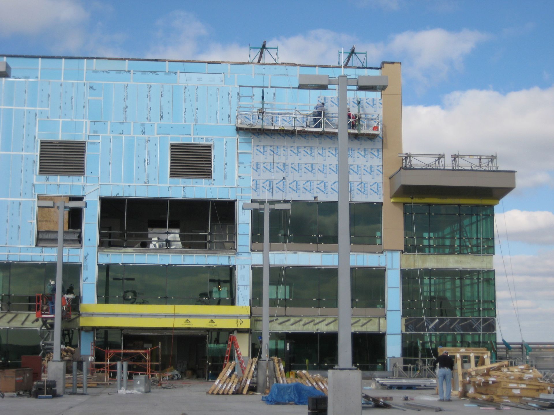 Building under construction with blue insulation panels, workers on suspended platform, clear windows.