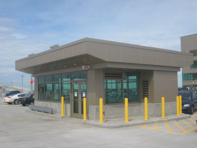 Tan security booth with glass windows and yellow bollards. Cars and a building are in the background.