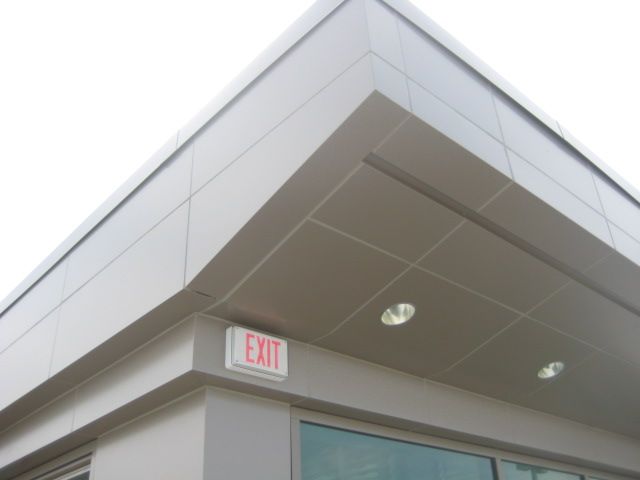 Exterior corner of a building with a light-colored metal facade, an exit sign, and recessed lighting.