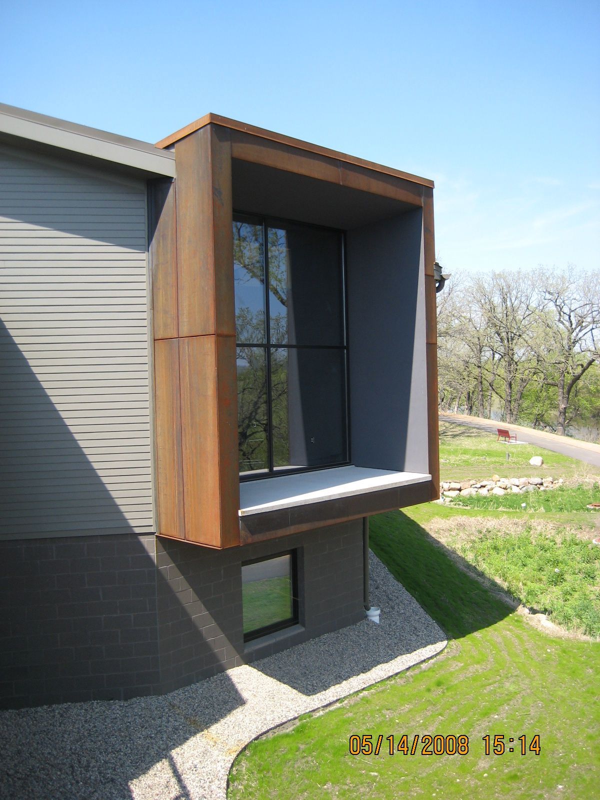 Modern house with large, boxy window frame, brown exterior, and gray siding. Sunny day with trees in the background.