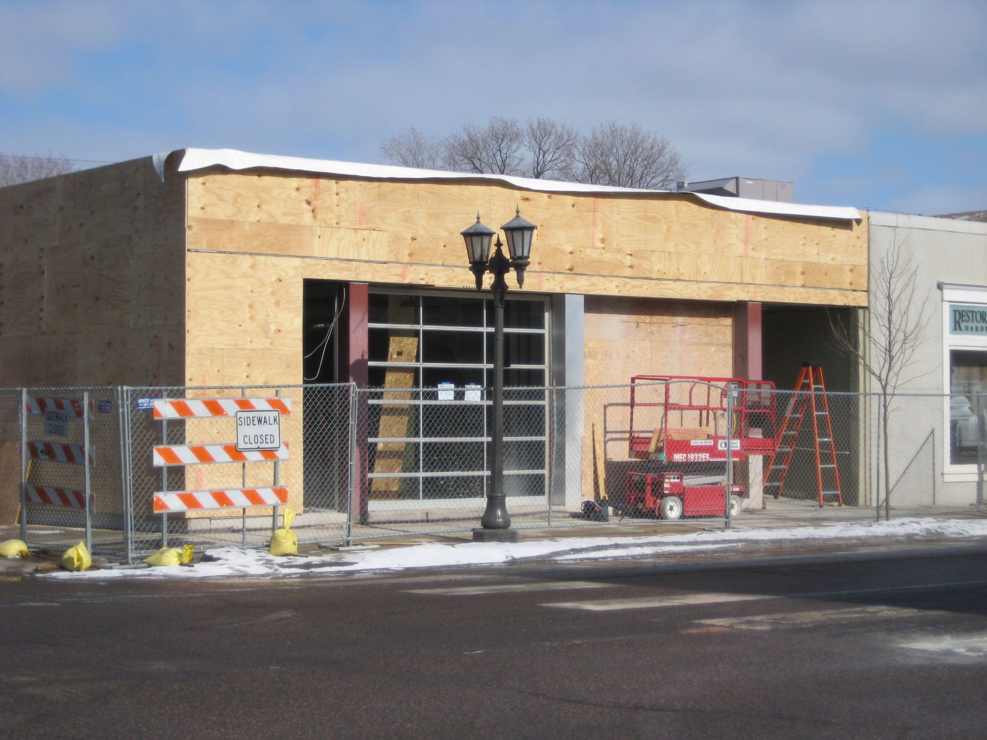 Building under construction, plywood siding, glass garage-style door, red lift, fenced off with snow-covered ground.