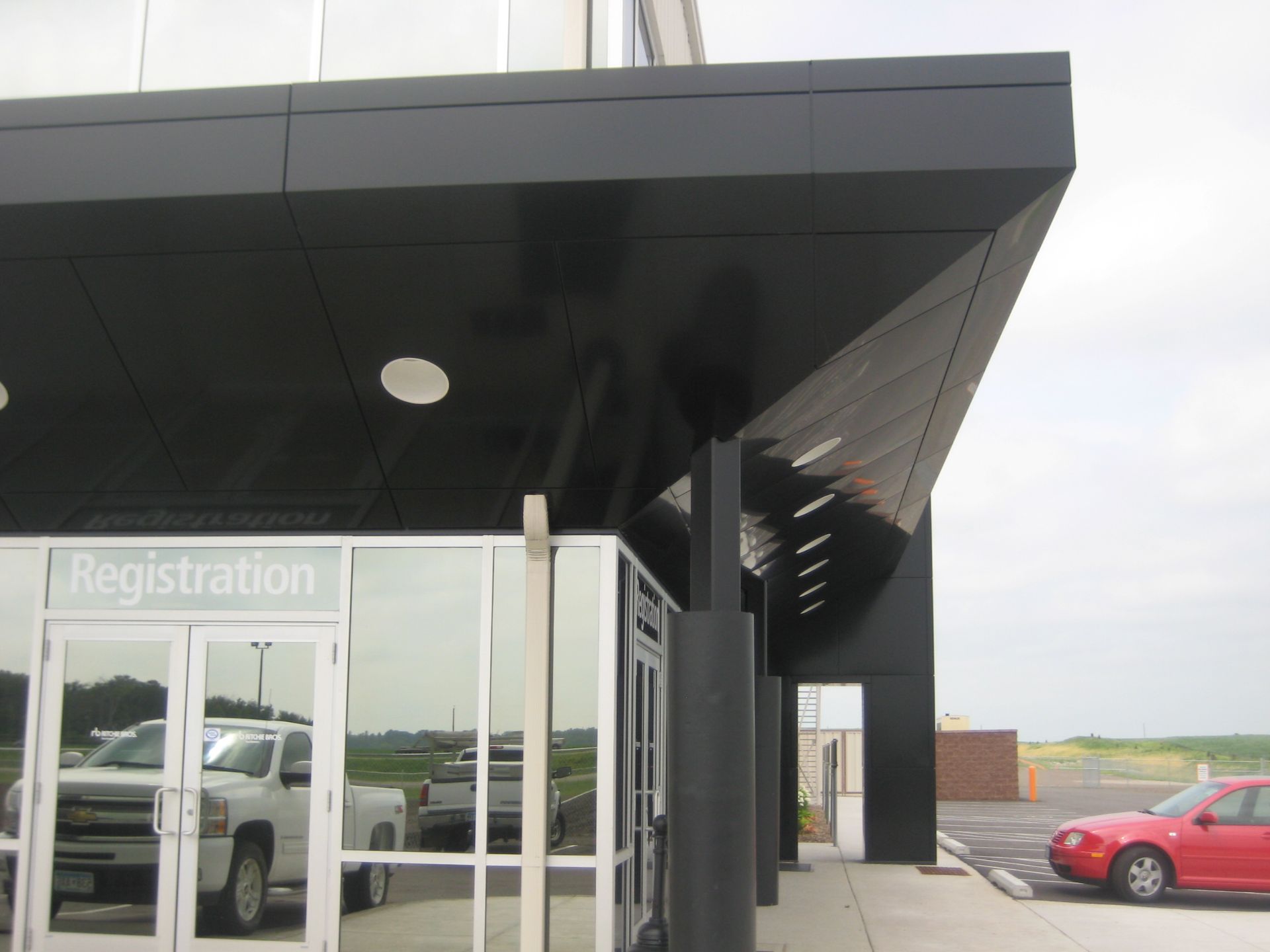 Black awning over a building's registration entrance with glass doors, a silver truck visible, and a red car parked.