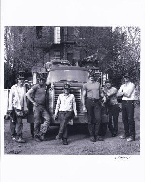 Group of six people posing in front of a truck, possibly construction workers. Old building in background.