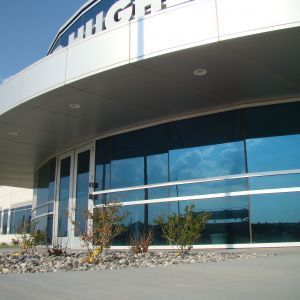 Modern building with curved roof, blue-tinted windows reflecting sky, small bushes in front.