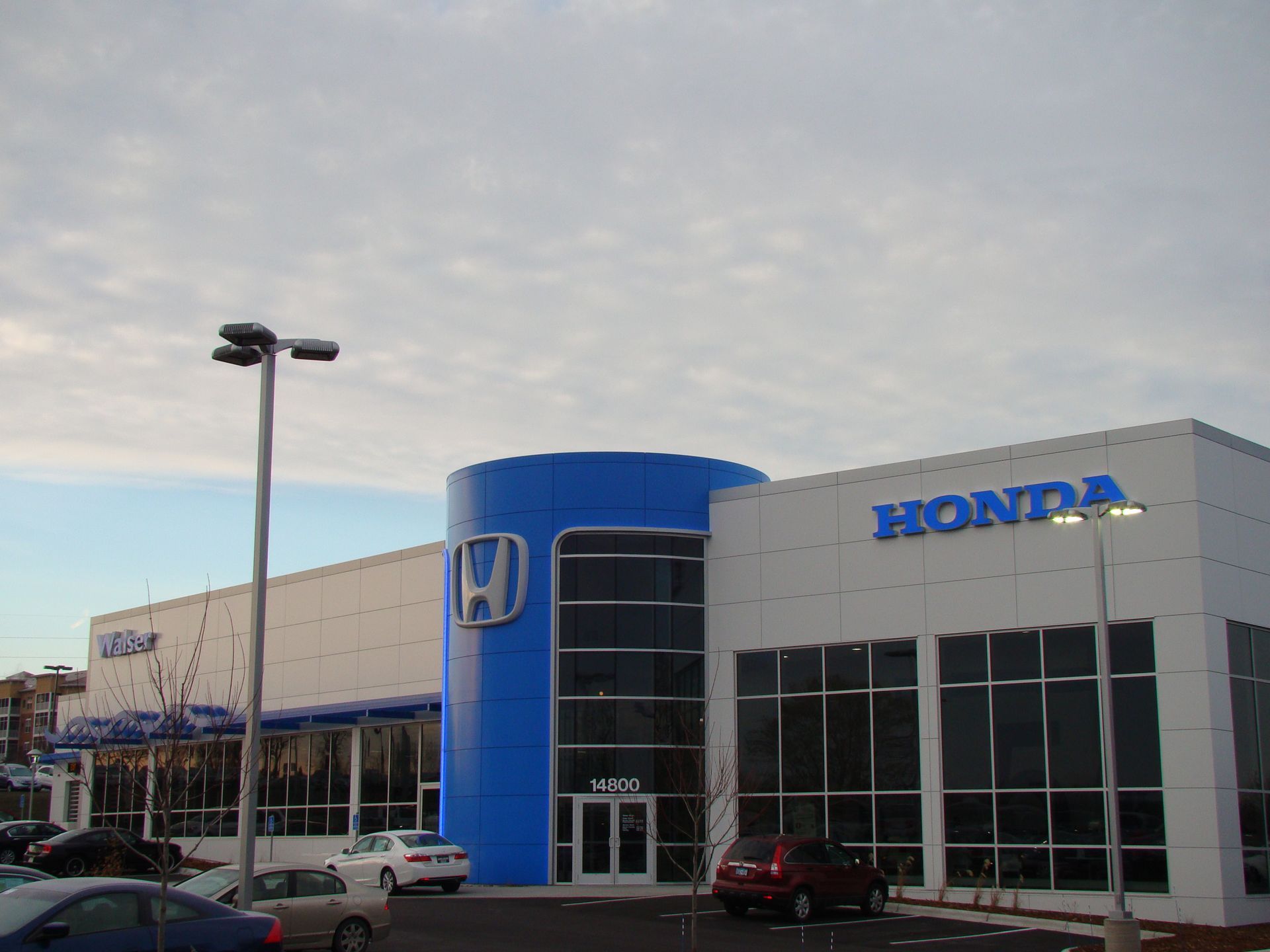 Honda dealership exterior with blue and white building, sign with Honda logo, and cars in the parking lot.
