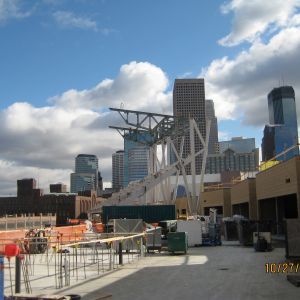 Construction site with a cityscape backdrop. Steel beams, equipment, and city buildings under a cloudy sky.