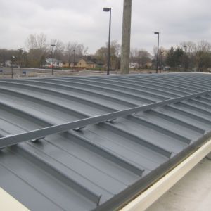 Gray metal roof with curved ridges and support beams, outdoors with cloudy sky and trees.