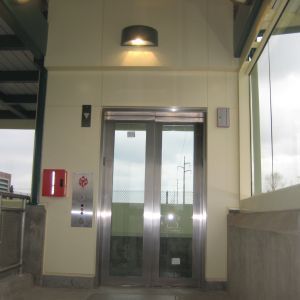 Elevator with glass doors at a public transit station. Controls, fire alarm box, and overhead light are visible.