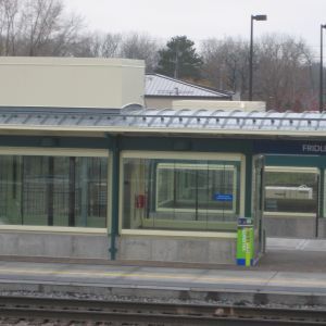 Train station platform with glass enclosures, green trim, and a roof, under cloudy skies.