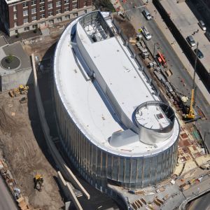 Aerial view of a modern building with a curved glass facade under construction, surrounded by construction equipment.
