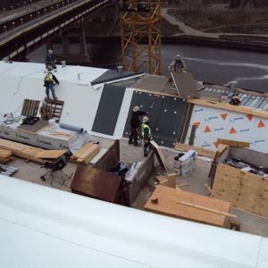 Construction workers on a rooftop, assembling materials near a crane, bridge in the background.