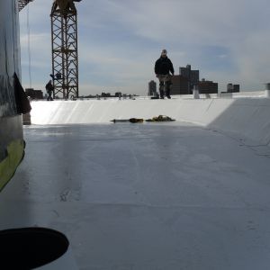 Workers on a flat white roof with a crane in the background; a clear sky.