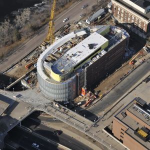 Aerial view of a building under construction, curved glass facade, next to a highway and bridge.