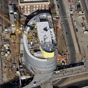 Construction site of a curved, modern building with a crane, road, and other buildings nearby.