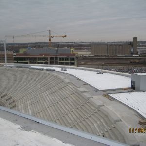 Construction site with large, curved roof. Crane in the background. Overcast sky.