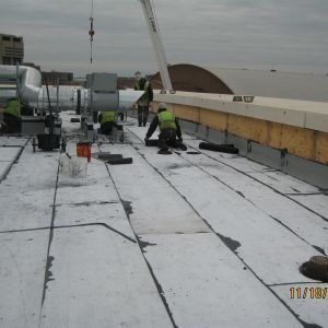 Workers on a rooftop install new roofing. A crane is present. Grey sky and city buildings are in the background.
