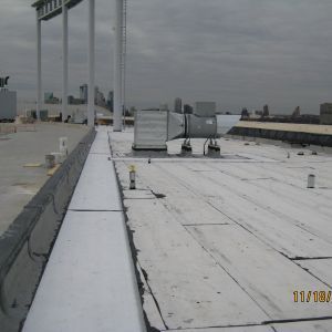 A flat commercial roof with HVAC equipment, metal posts, and a city skyline under a cloudy sky.