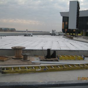 Construction site rooftop, white membrane laid, tools and ladder in foreground, building in the background, cloudy sky.