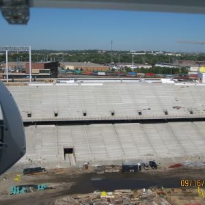 Construction of a stadium with concrete tiers. Blue sky, distant buildings, and construction materials visible.