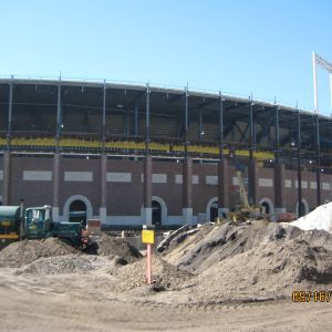 Stadium exterior under construction with dirt piles, machinery, and a clear blue sky.