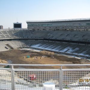 Empty football stadium, dirt field, snowy seating, a digital scoreboard, and a protective railing.