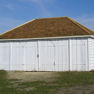 White building with brown shingled roof, three large doors, set against a blue sky and grassy ground.