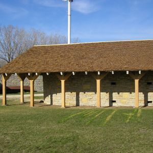 Stone building with wooden posts and a brown shingle roof on a grassy field, with a tall white pole visible.