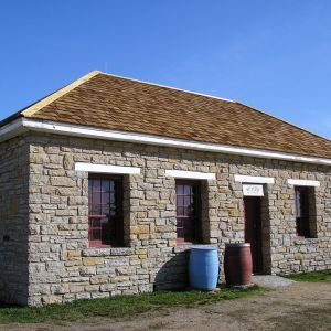 Stone building with brown roof, blue barrel, and brown barrel in front, on a sunny day.