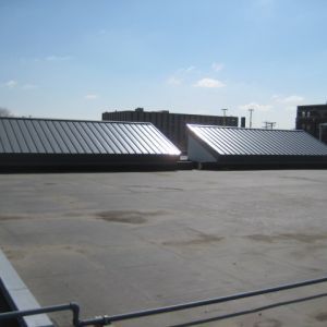 Flat rooftop with two metal-clad rectangular structures; buildings in background under a blue sky.