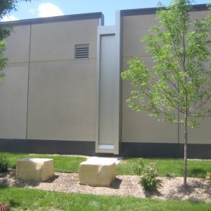 Exterior of a building with a tall, narrow silver structure and two stone blocks on the grass.
