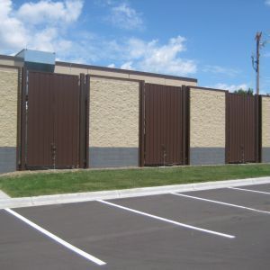 Brown metal fence segments against tan and gray building exterior; asphalt parking lot.