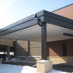 Black awning over an entrance supported by a dark column on a snowy day. Brick building in background.