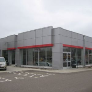 Car dealership building with silver and red accents, large windows. Cars are visible inside. Cloudy sky.