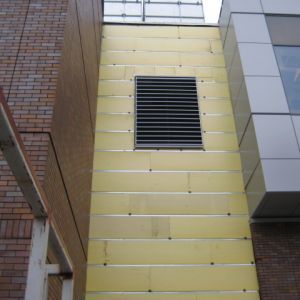 Yellow-paneled exterior wall with a dark vent and adjacent brick and glass sections.
