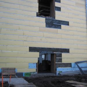 Exterior wall of a building under construction, showing insulation panels and openings for a door and window.