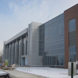 Modern building with glass windows, silver panels, and brick facade; bike rack in front, snow on the ground.