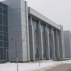 Modern gray building with large windows and vertical columns, snow on the ground, trees in front.