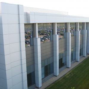 Modern building with glass windows, gray and white facade, and a parking lot visible in the windows.