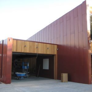 Modern building with reddish-brown metal siding and wooden entryway. Blue wheelbarrow inside open garage.