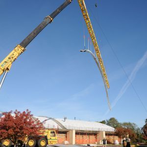 Crane lifting a long, curved metal beam over a building under construction on a sunny day.