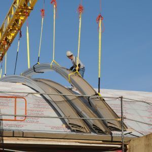 Construction worker installing curved metal roof panels, lifted by crane.