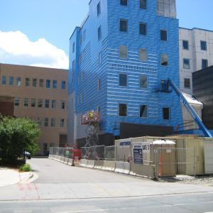 Building under construction; blue exterior cladding, construction workers on lift, safety barriers, and porta-potty visible.