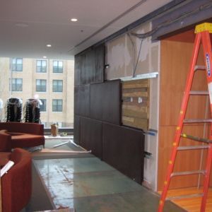 Interior under construction: brown wall panels, orange ladder, building view through windows, partially finished floor.