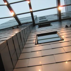Modern building exterior with shiny metal paneling, small windows, and glass ceiling.
