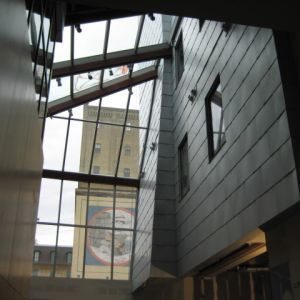 Interior view of a modern building with large windows. A tall building with a sign is visible outside.