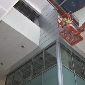 Worker in lift installing panels on building's corner with glass windows below.
