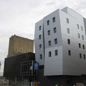 Modern metallic building with a curved, angular design next to a historic tan building.