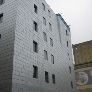 Gray rectangular building with evenly spaced windows, next to a brown building with a painted mural.