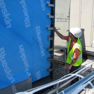 Construction worker in hard hat and vest installs blue Blueskin membrane on building exterior.