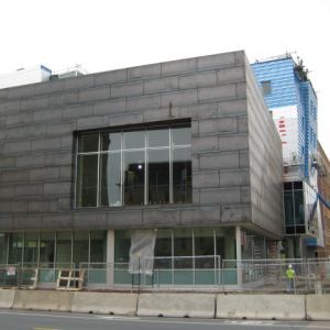 Exterior of a building under construction. Grey stone façade with large window. Construction barriers and workers present.